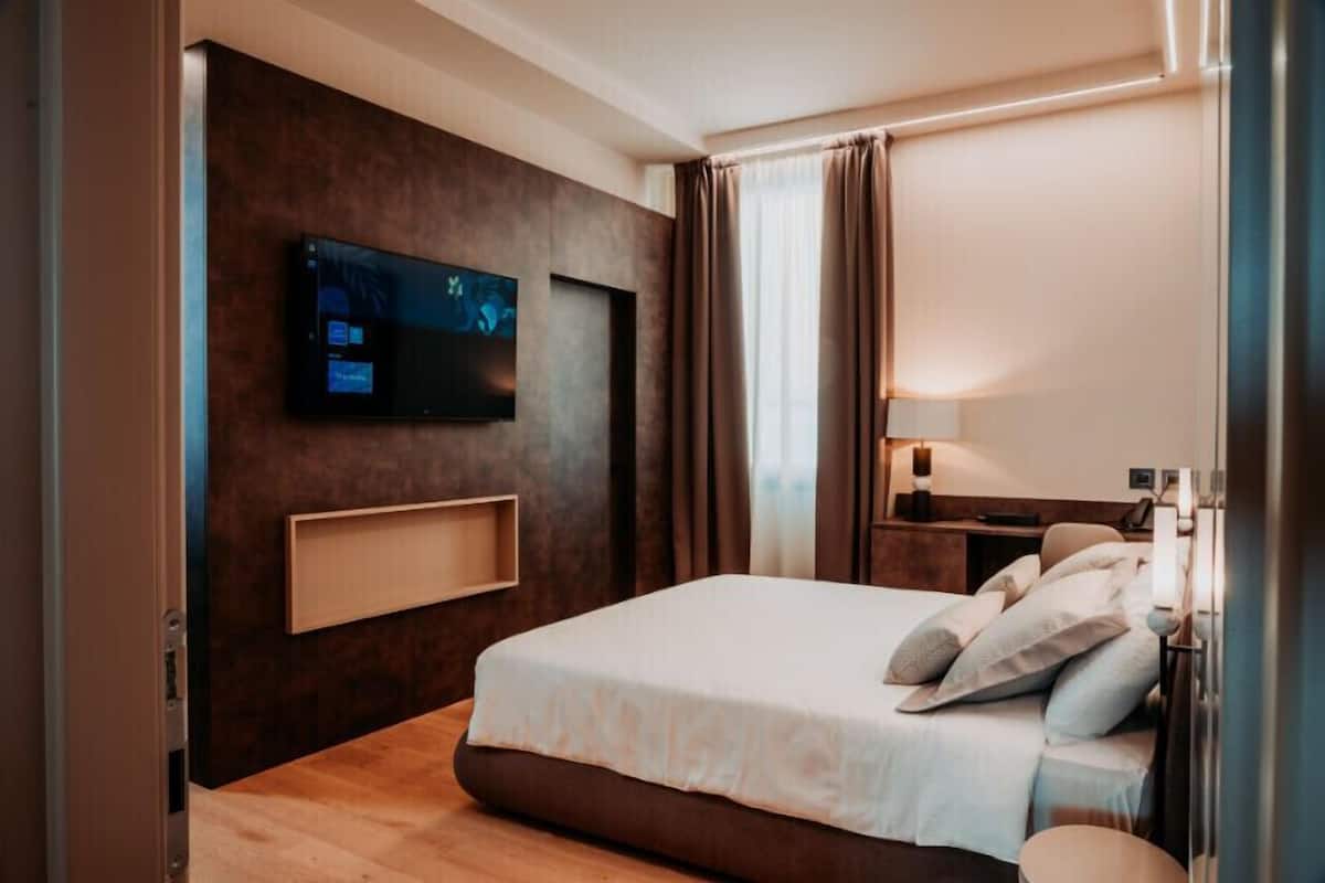 A modern hotel room with a large bed, a flat-screen TV mounted on the wall, and a bedside table with a lamp.