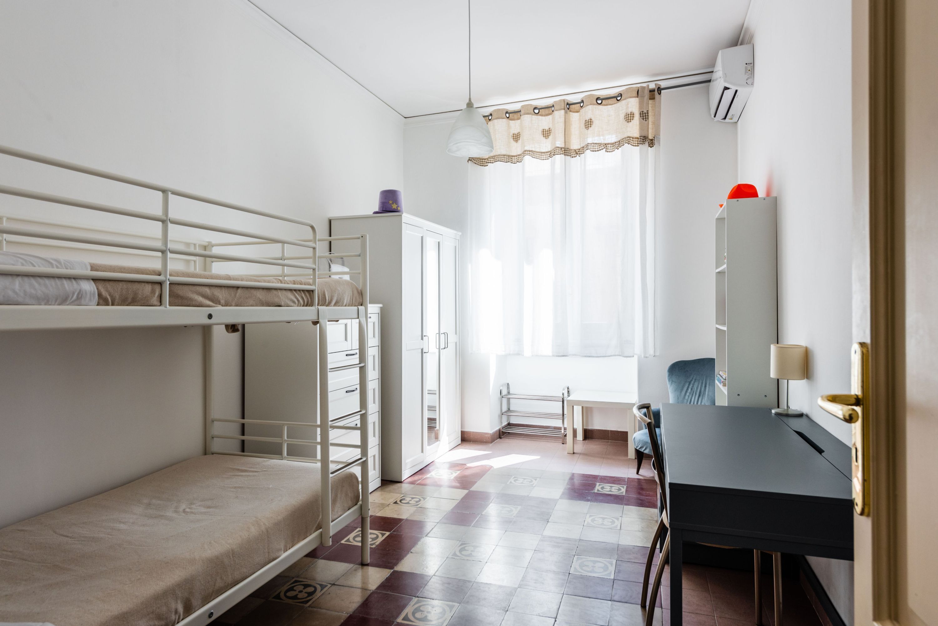 A dormitory room with bunk beds, a desk, a chair, a wardrobe, and a window with curtains.