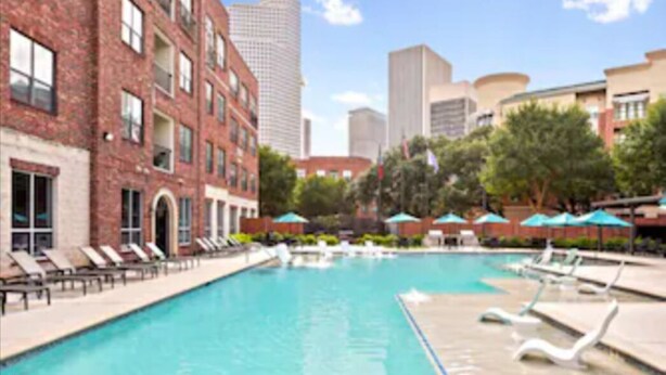 A swimming pool with lounge chairs and umbrellas, surrounded by brick buildings and modern high-rises. -  New! Luxury Midtown Escape (Houston)