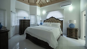 A bedroom with a large bed, wooden headboard, and a ceiling with wooden beams. There are two windows with blinds, a dresser, and a nightstand with a lamp. - Island Dreams Villa at Legacy Townhouse (Montego Bay)