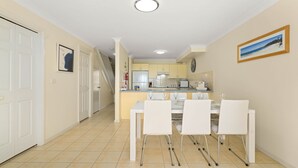 A modern kitchen with white cabinets, a dining area with a white table and chairs, and a hallway with a white door. - Pier 8 by Experience Jervis Bay (Huskisson)