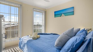 A bedroom with a bed, pillows, and a painting of a beach scene. - Pier 8 by Experience Jervis Bay (Huskisson)