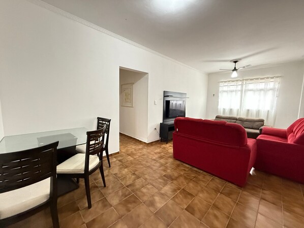 A living room with a glass dining table, two chairs, a red sofa, a TV, and a ceiling fan. - Pé na Areia Praia da Guilhermina II (Praia Grande)