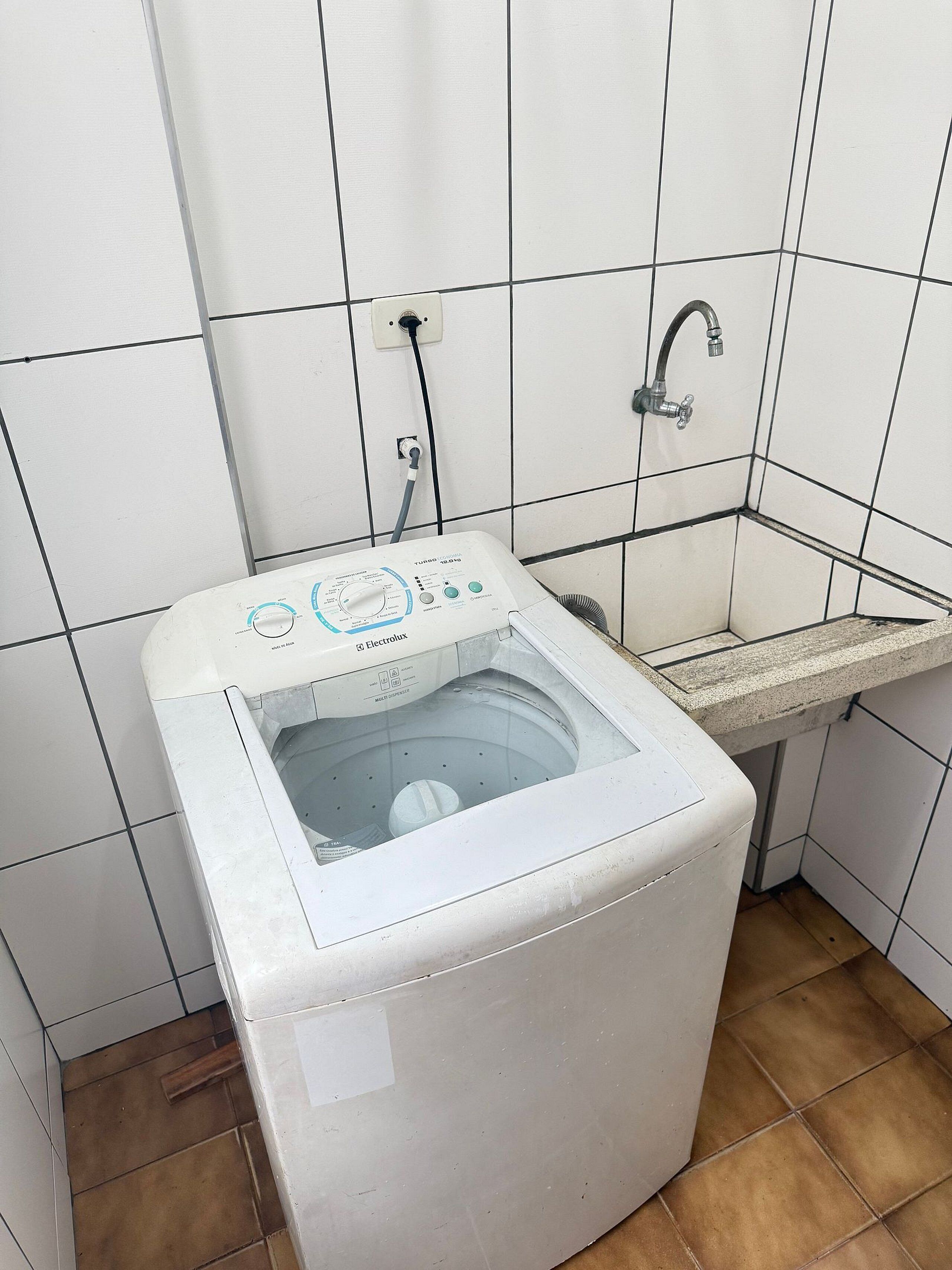 A compact, white washing machine in a small bathroom with white tiled walls and a single sink.