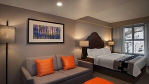 Une chambre d’hôtel avec un canapé gris, des coussins orange, un lit recouvert d’une couette blanche, une tête de lit en bois et une fenêtre donnant sur un paysage enneigé.
