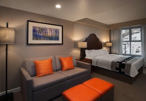 Quarto de hotel com sofá cinzento, almofadas laranja, cama com edredom branco, cabeceira de madeira e janela com vista para uma paisagem nevada.