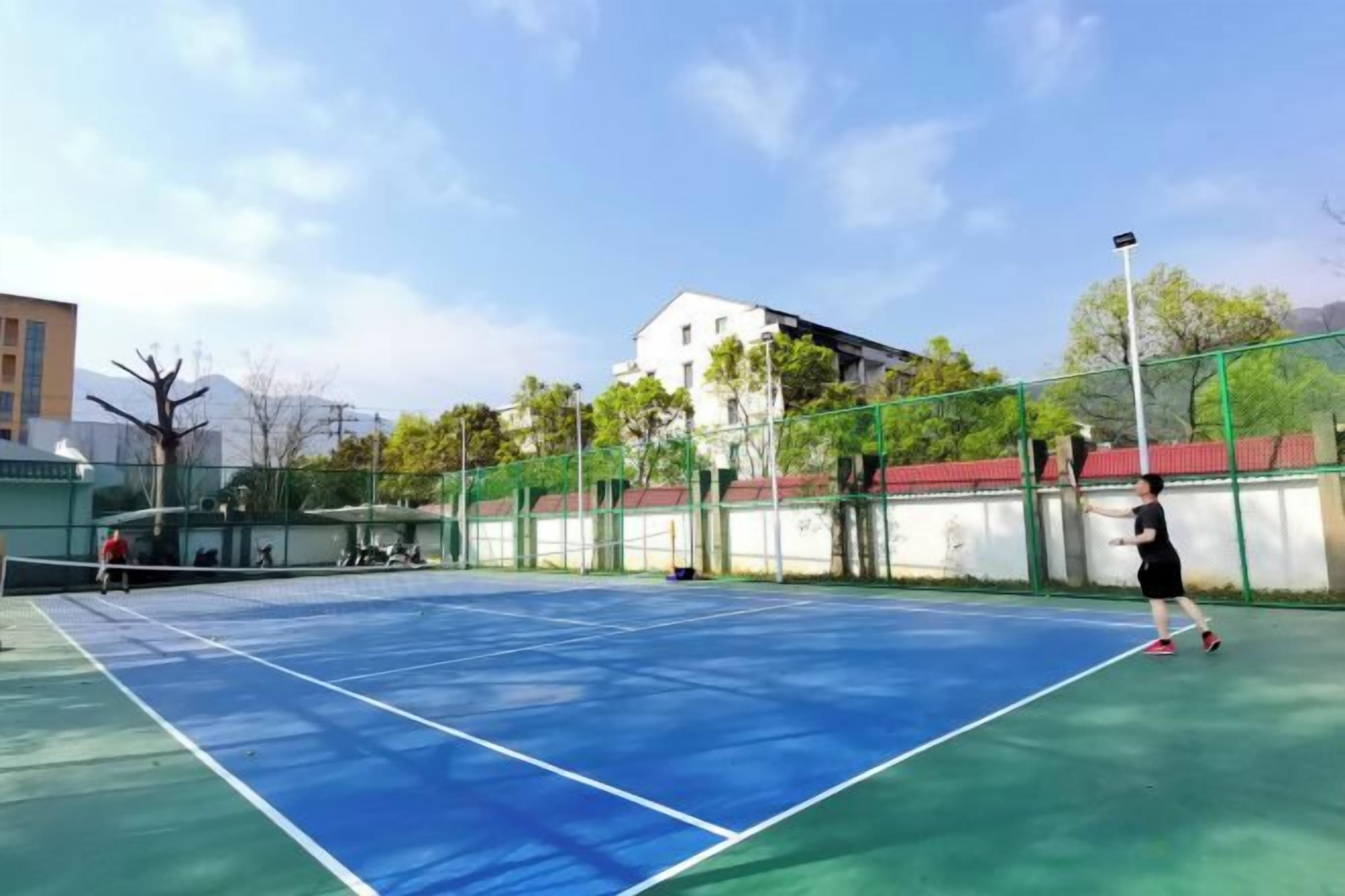 Una cancha de tenis al aire libre con red, rodeada por una cerca verde, y edificios al fondo.