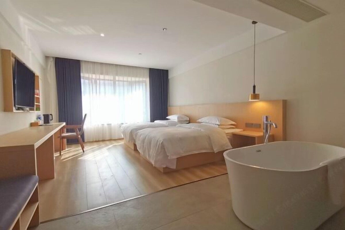 A modern hotel room with a large bed, a sitting area with a desk, a large bathtub, and a large window with curtains.