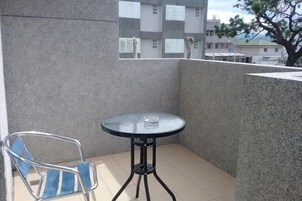 Un balcón con una mesa redonda de vidrio, una silla, piso de baldosas y vistas a edificios y un árbol.