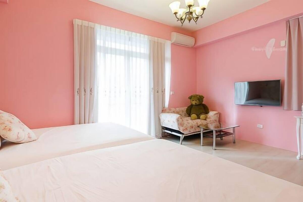A pink-themed bedroom with two beds, a chair with a teddy bear, a TV, and an air conditioning unit.