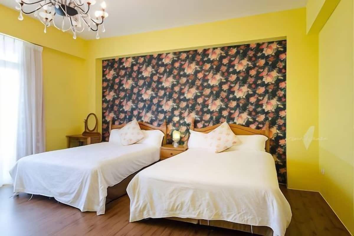 A hotel room with two beds, a floral headboard, a chandelier, and a bedside table with a lamp.