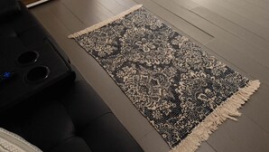 Ein Zimmer mit einem schwarzen Ledersofa, einem gemusterten Teppich, einem weißen Stehlampen-Spiegel und einer Holzkabine mit Schubladen.