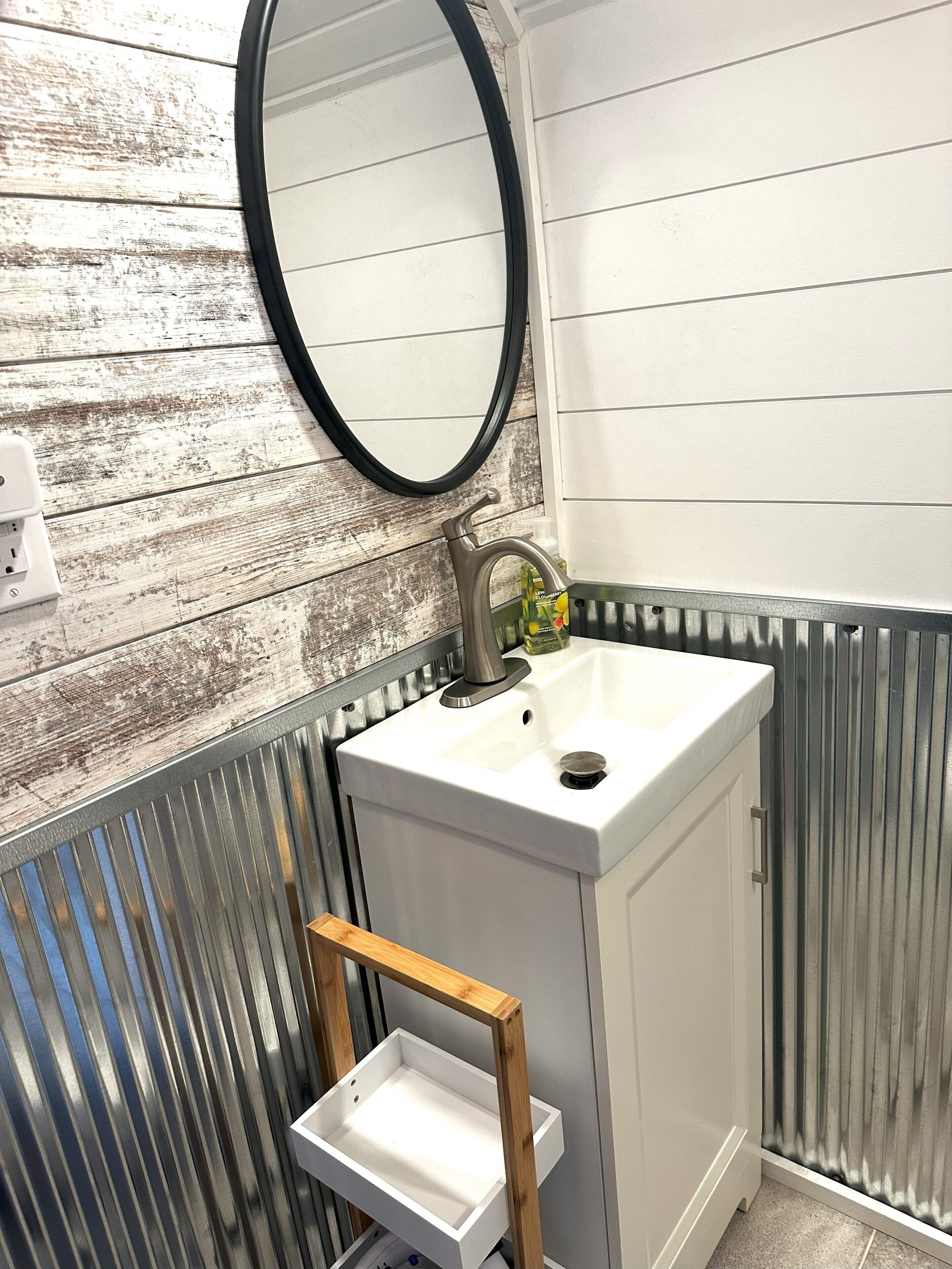 Un baño con un lavamanos blanco, un espejo redondo y una repisa de madera.