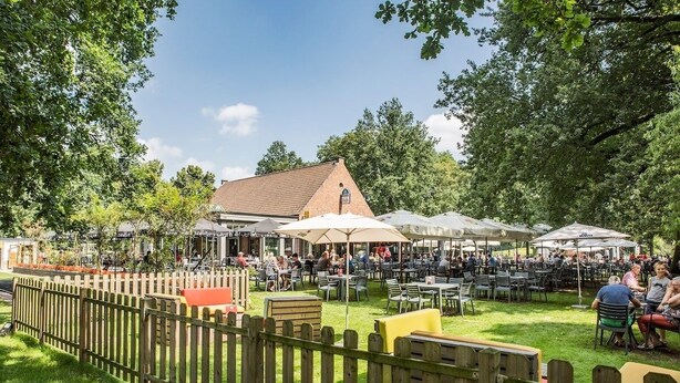 An outdoor dining area with tables and umbrellas, surrounded by a wooden fence and greenery. - Familyfriendly house for 12 persons (Kasterlee)