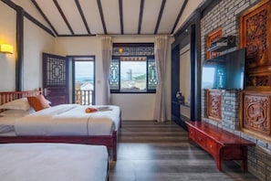A hotel room with two beds, a TV, a wooden bench, and a view of the sea through a window.