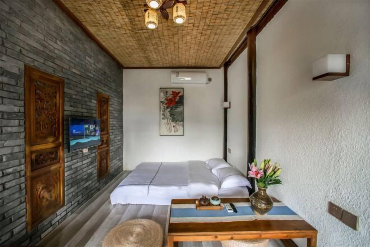 A bedroom with a bed, a television, a wall-mounted air conditioner, and a small table with a vase of flowers.
