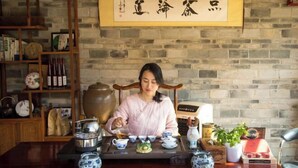 A person seated at a table with traditional Chinese tea set and utensils, a wooden bookshelf with various items, and a stone wall with a framed calligraphy piece.