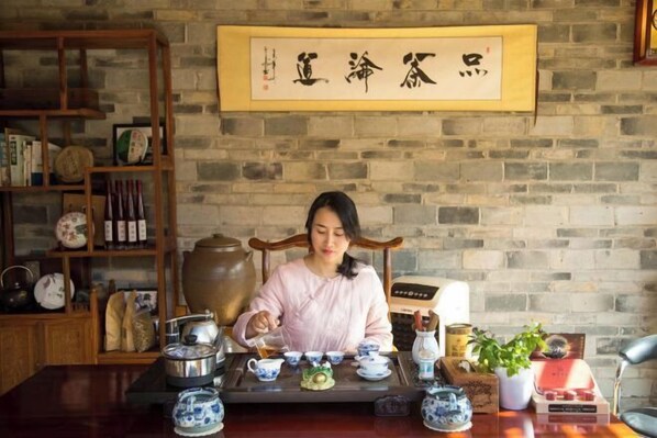 A person seated at a table with traditional Chinese tea set and utensils, a wooden bookshelf with various items, and a stone wall with a framed calligraphy piece. - Colorful·Muyun inn (Tengchong)