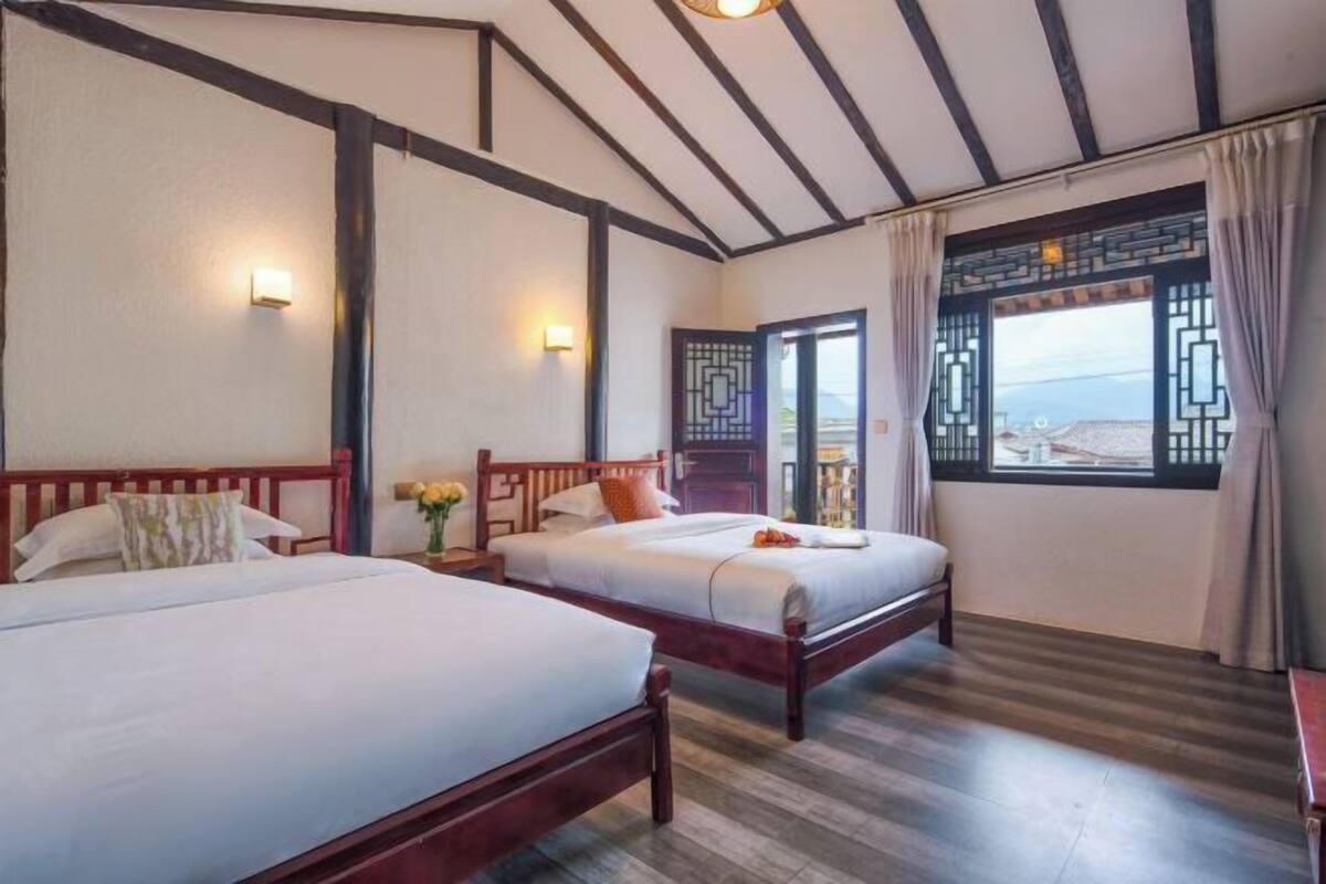 A hotel room with two beds, wooden beams on the ceiling, a large window with curtains, and wall-mounted lights.