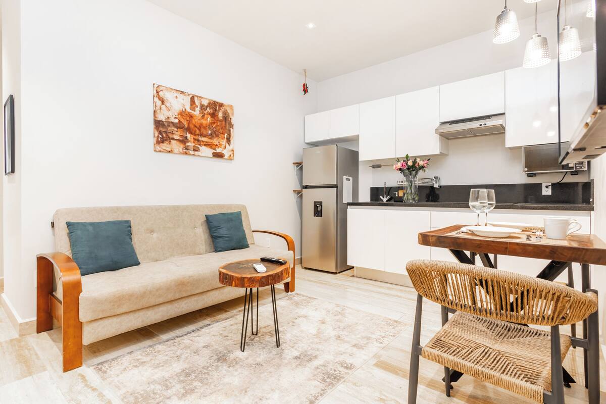 Ein modernes Wohnzimmer mit einer Couch, einem Couchtisch und einem Korbstuhl. Im Hintergrund ist eine Küche mit weißen Schränken und Edelstahlgeräten zu sehen.