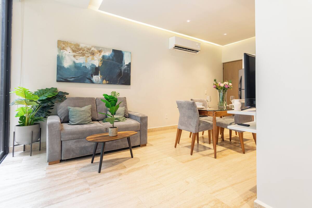 Ein modernes Wohnzimmer mit grauem Sofa, Couchtisch und Essbereich mit Stühlen und Tisch. An der Wand hängt ein großes abstraktes Gemälde und auf dem Sofa steht ein Blumentopf.