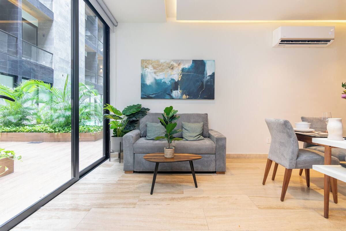 Ein modernes Wohnzimmer mit grauem Sofa, einem Holztisch und einem Essbereich mit weißem Tisch und grauen Stühlen. Ein großes Fenster bietet Blick auf Grünflächen.