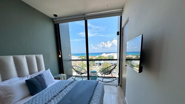A bedroom with a large bed, a TV mounted on the wall, and a balcony with chairs. - Musa del Carmen By BoutiqueApartments MX (Playa del Carmen)