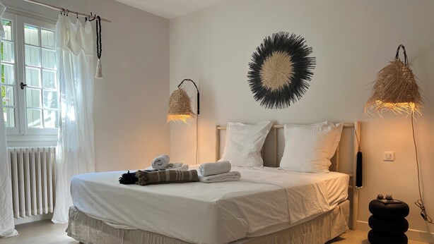 A neatly made bed with white linens and a beige skirt, a round wall decoration, and two wall-mounted lamps with natural materials. - Fantastic private villa with heated saltwater pool in Provence (Modène)