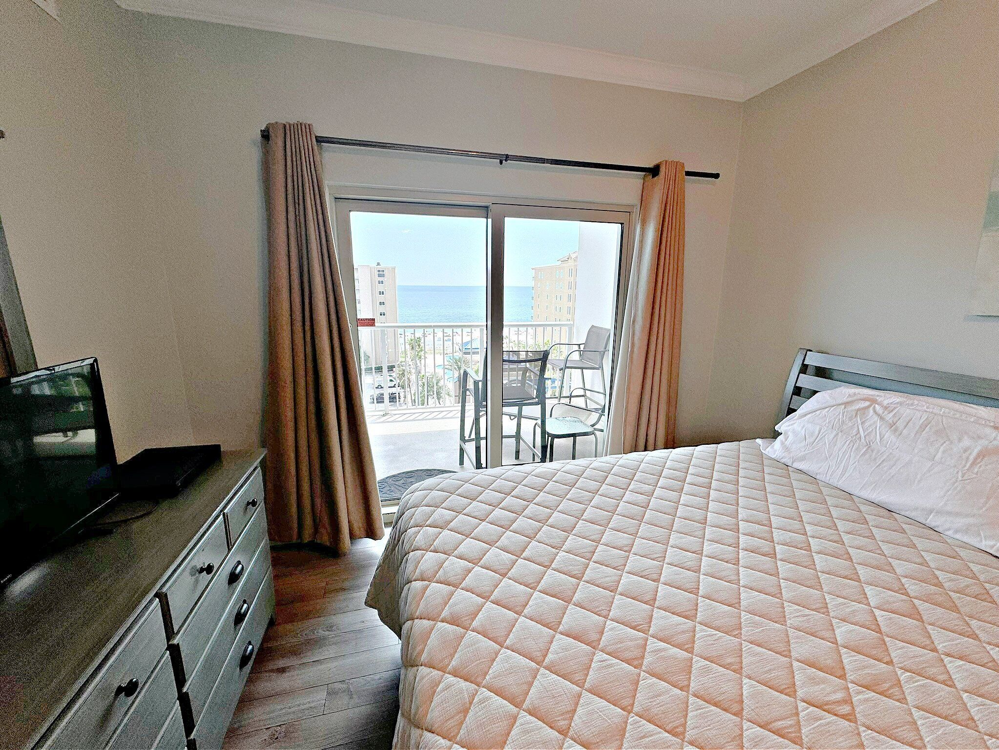 A hotel room with a bed, a dresser, a TV, and a balcony with a view of the sea and buildings.