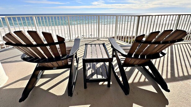 Varanda com duas cadeiras Adirondack e uma pequena mesa, com vista para uma praia e um corrimão branco.