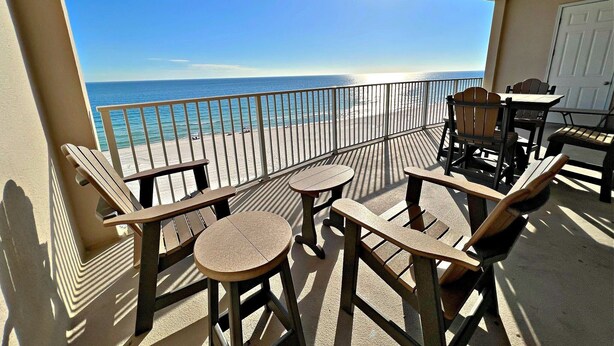 A balcony with a view of the beach and ocean. There are wooden chairs and tables, and a door is visible in the background. - Regency Isle 703 (Orange Beach)