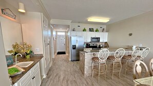 A modern kitchen with white cabinets, a stainless steel refrigerator, a microwave, and a dining area with white chairs. - Regency Isle 703 (Orange Beach)