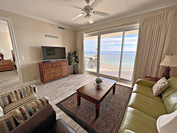 A living room with a television, a sofa, a coffee table, and a view of the ocean through large windows. - Regency Isle 704 (Orange Beach)