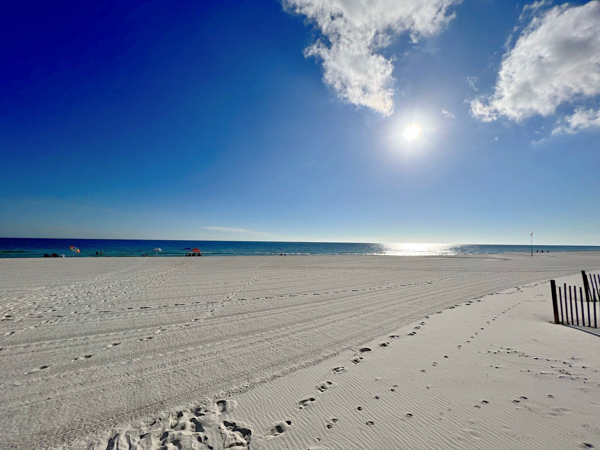 Une vaste plage de sable fin, parsemée d’empreintes menant à l’eau, un ciel dégagé et un soleil éclatant.