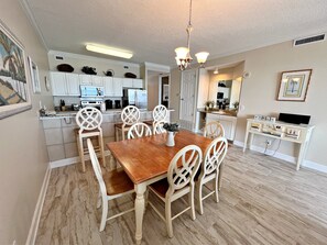 A dining area with a wooden table and chairs, a kitchen in the background, and a small TV on a white cabinet. - Regency Isle 704 (Orange Beach)