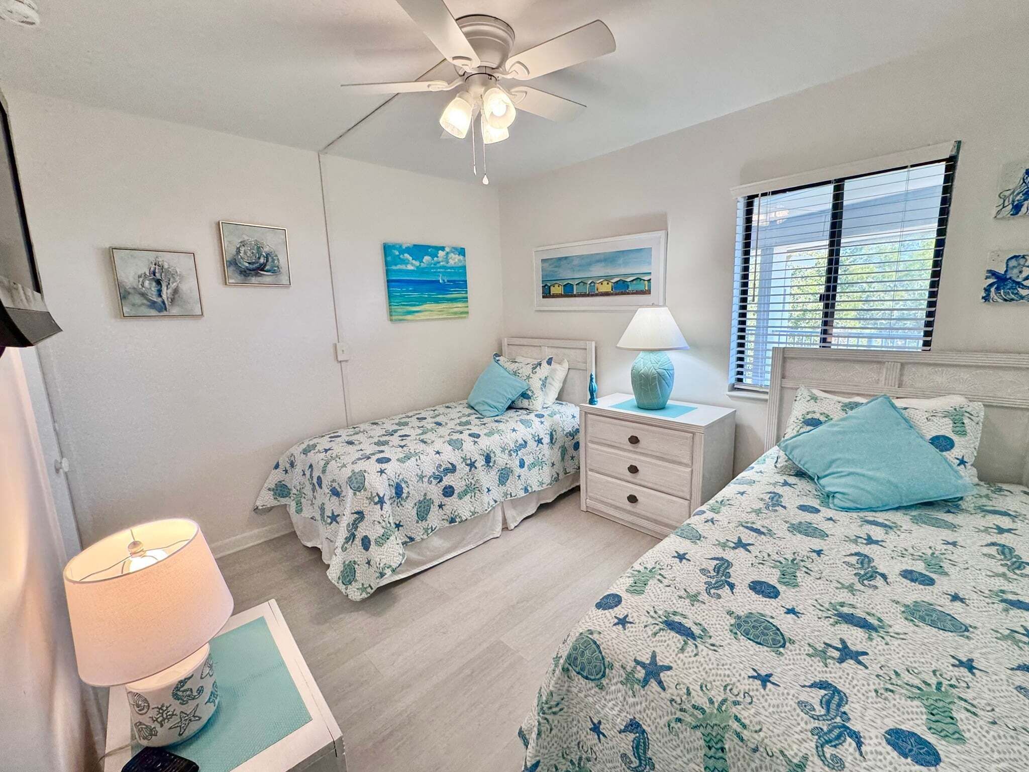 A bedroom with two beds, a ceiling fan, artwork on the walls, and a window with blinds.