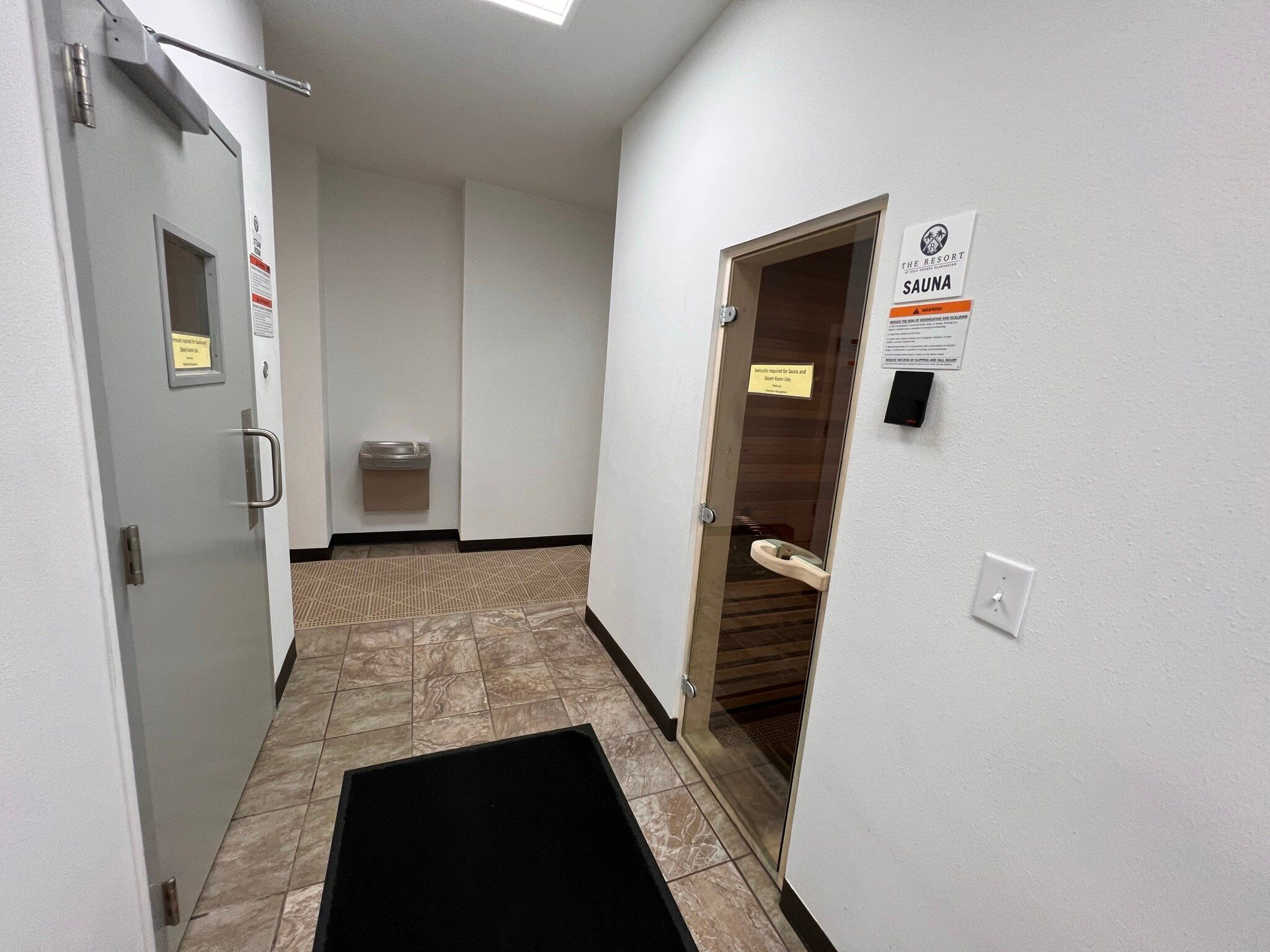 Pasillo con puerta de sauna, dispensador de desinfectante para manos y un letrero que indica que el uso de la sauna es obligatorio para acceder a la sauna y al baño de vapor.