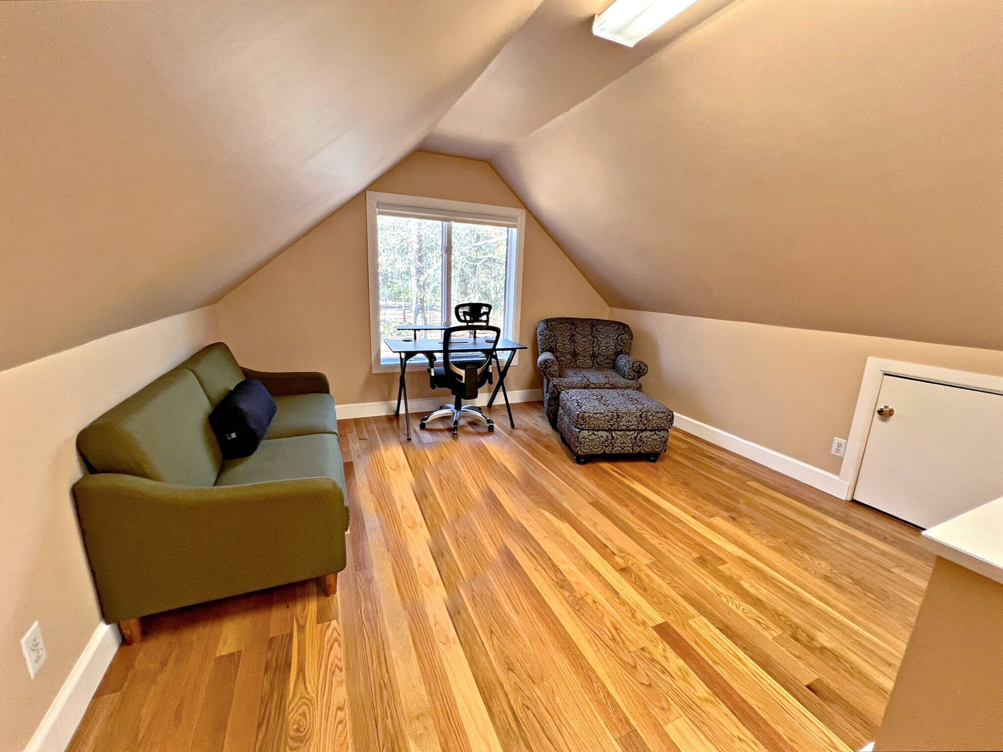 An attic bedroom with a wooden floor, a green sofa, a patterned armchair, and a desk with a chair.