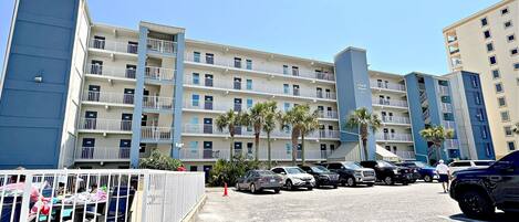 A multi-story hotel with balconies, a parking area, and palm trees.