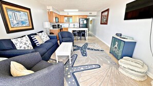 A living room with a sofa, coffee table, and a starfish-patterned rug. There is a kitchen in the background with wooden cabinets and a dining area. - Island Shores (Gulf Shores)