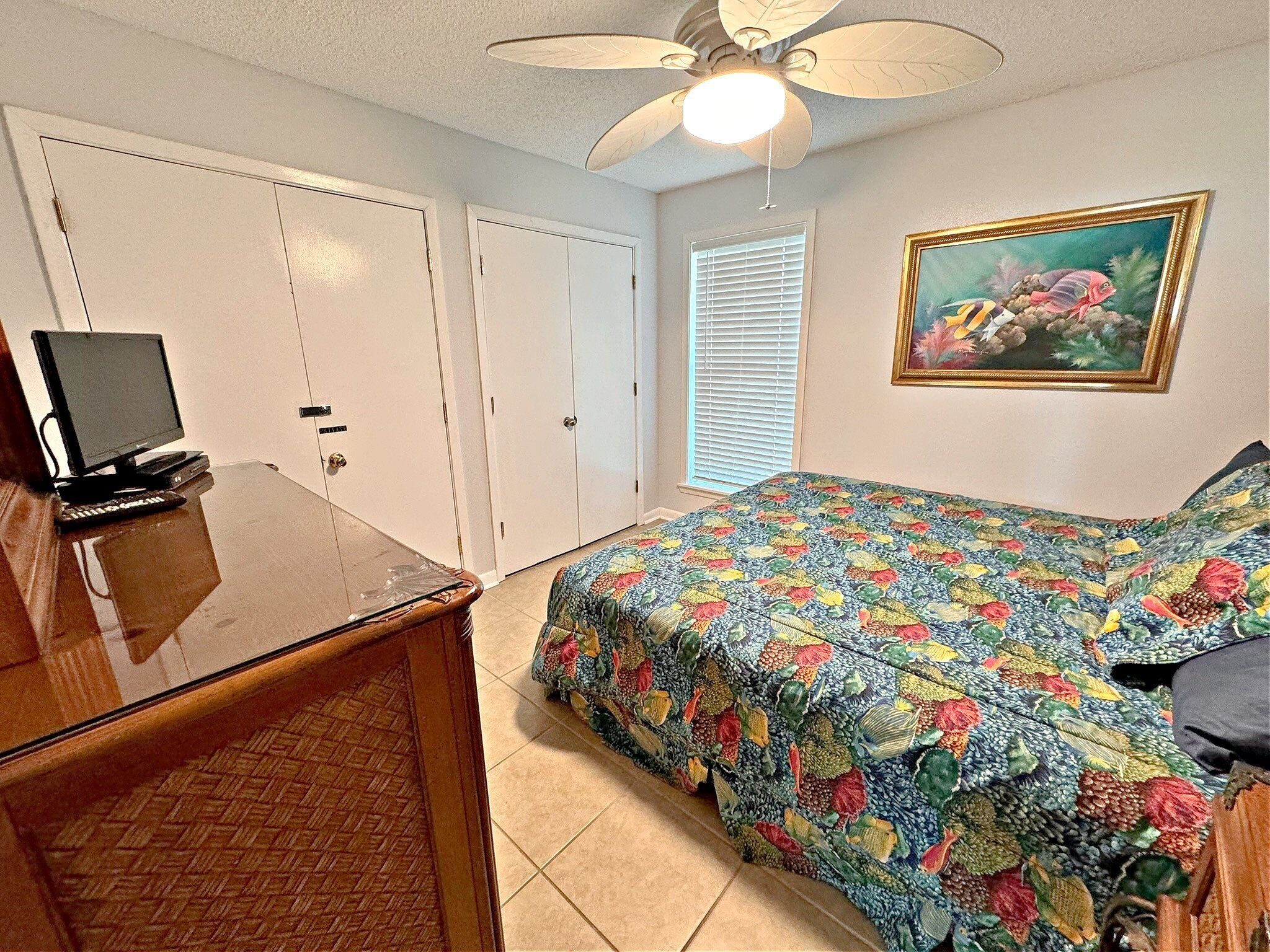 A bedroom with a bed, a desk with a computer, a ceiling fan, and a window with blinds.