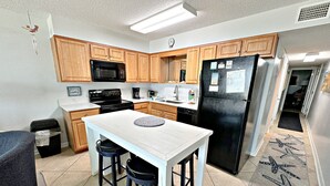 A kitchen with wooden cabinets, a black refrigerator, a stove, and a sink. There is a small island with stools and a clock on the wall. - Island Shores (Gulf Shores)
