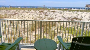 A beach view from a balcony with green chairs and a table, overlooking a sandy beach with sparse vegetation and a clear sky. - Island Shores (Gulf Shores)