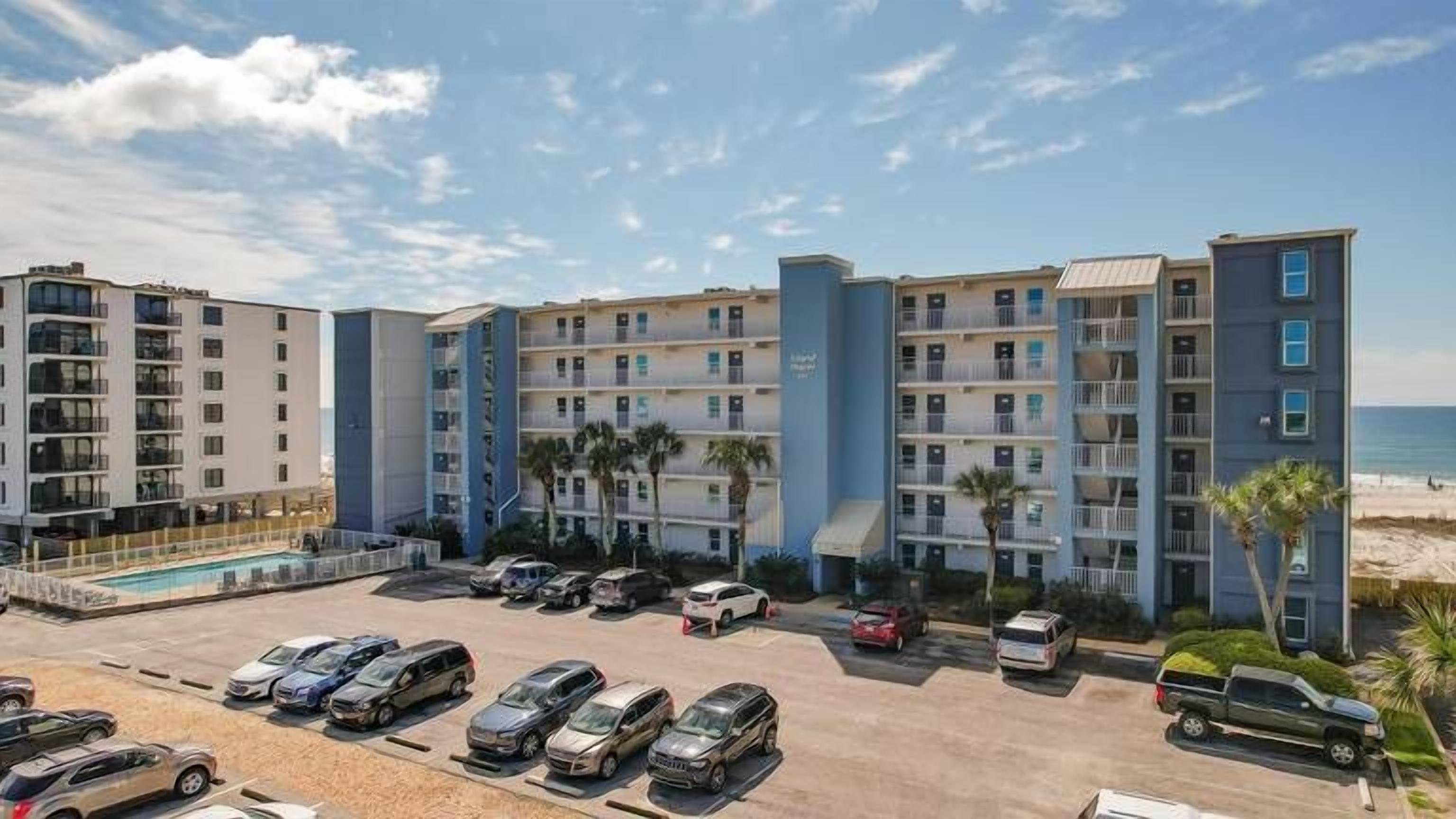 A multi-story hotel with a pool, parking lot, and beach view.