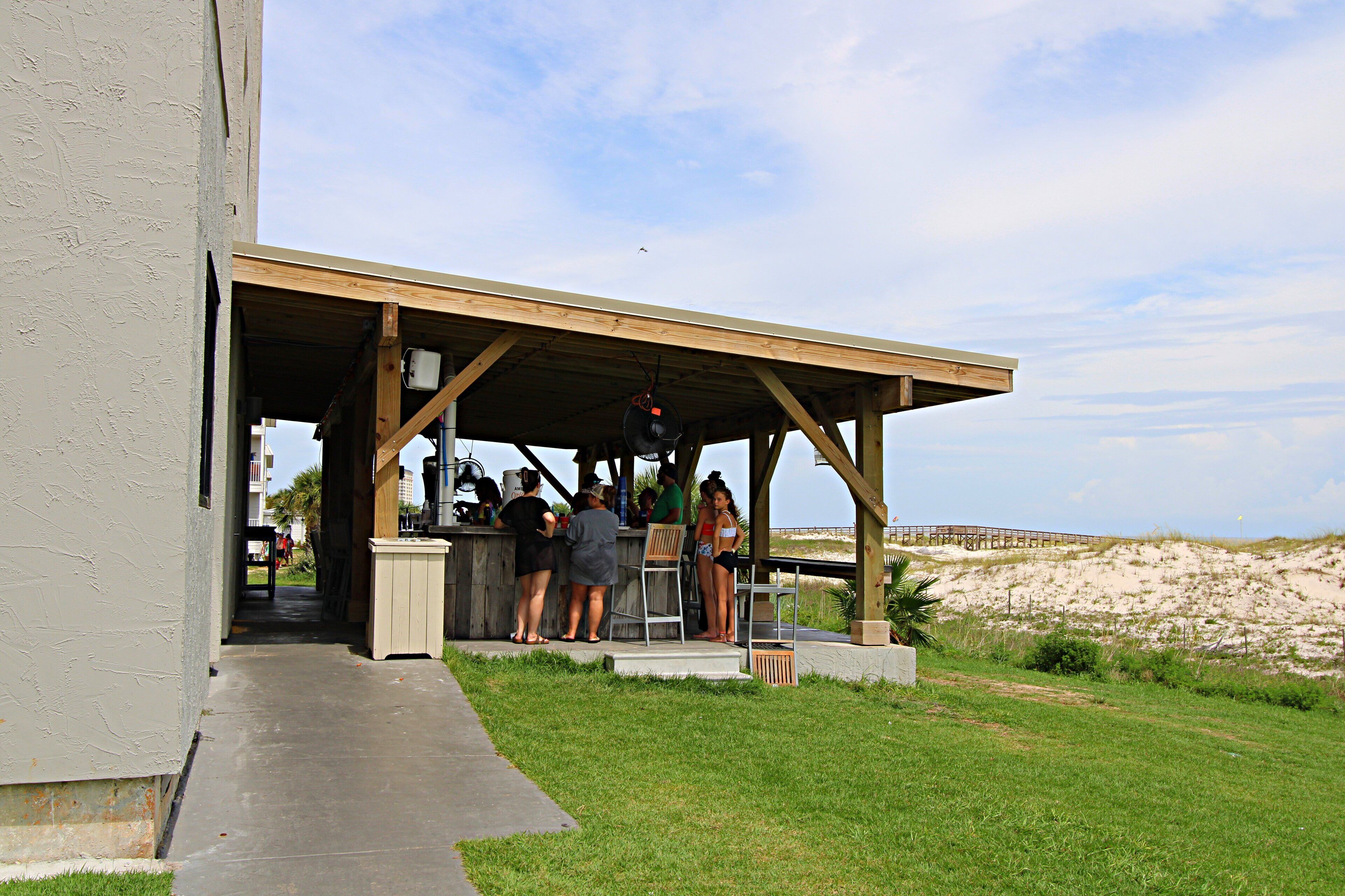A covered outdoor seating area with a concrete pathway leading to it, surrounded by grass and a sandy beach in the background.