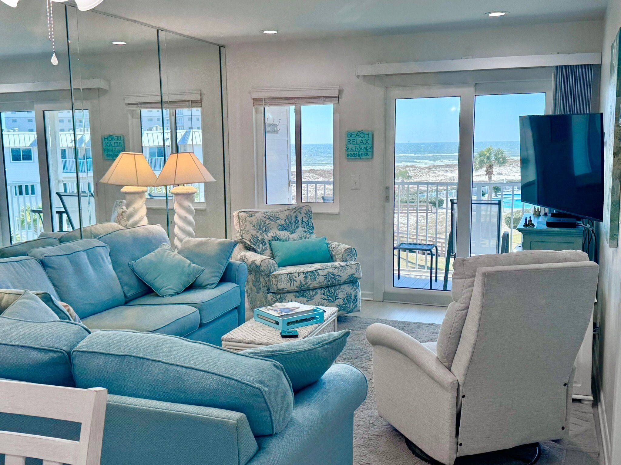 Una sala de estar con sofá, sillón y televisor. Se ven ventanas con vista a la playa.