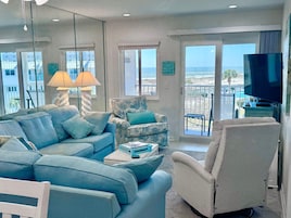 A living room with a sofa, armchair, and a television. There are windows with a view of the beach.