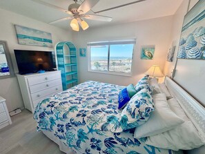 Une chambre à coucher comprenant un lit, une télévision, un ventilateur de plafond, une fenêtre avec vue et une décoration sur le thème de la plage.