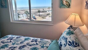 A bedroom with a large window overlooking a beach, a bed with floral bedding, a lamp, and a painting on the wall. - Gulf Shores Plantation (Gulf Shores)