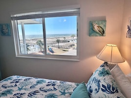 Un dormitorio con un ventanal grande con vistas a una playa, una cama con ropa de cama floral, una lámpara y un cuadro en la pared.